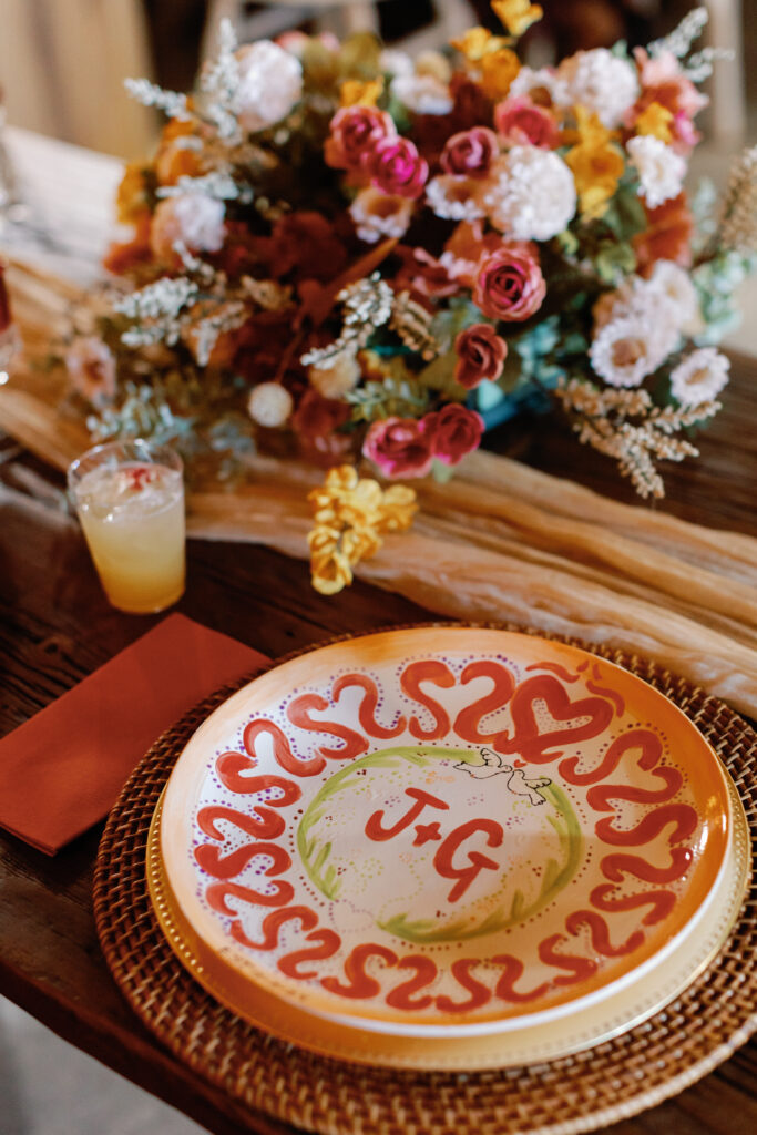 A bride and groom dinner plate designed and painted by the couple sits at the sweetheart table surrounded by colorful flowers