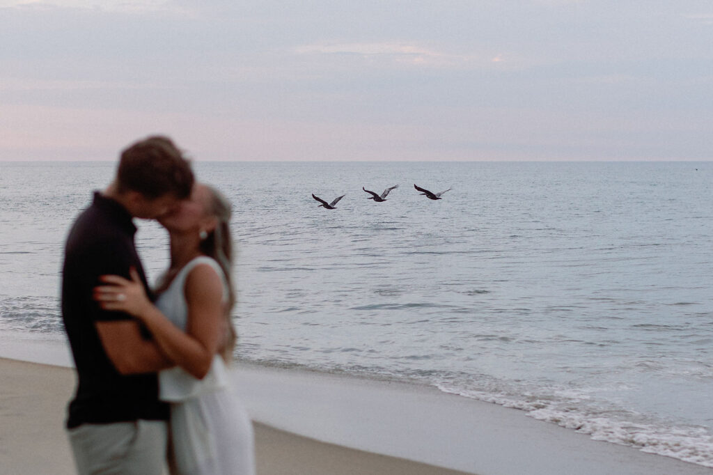 Couple celebrating after a surprise proposal with joyful embrace on a North Carolina beach at sunset.