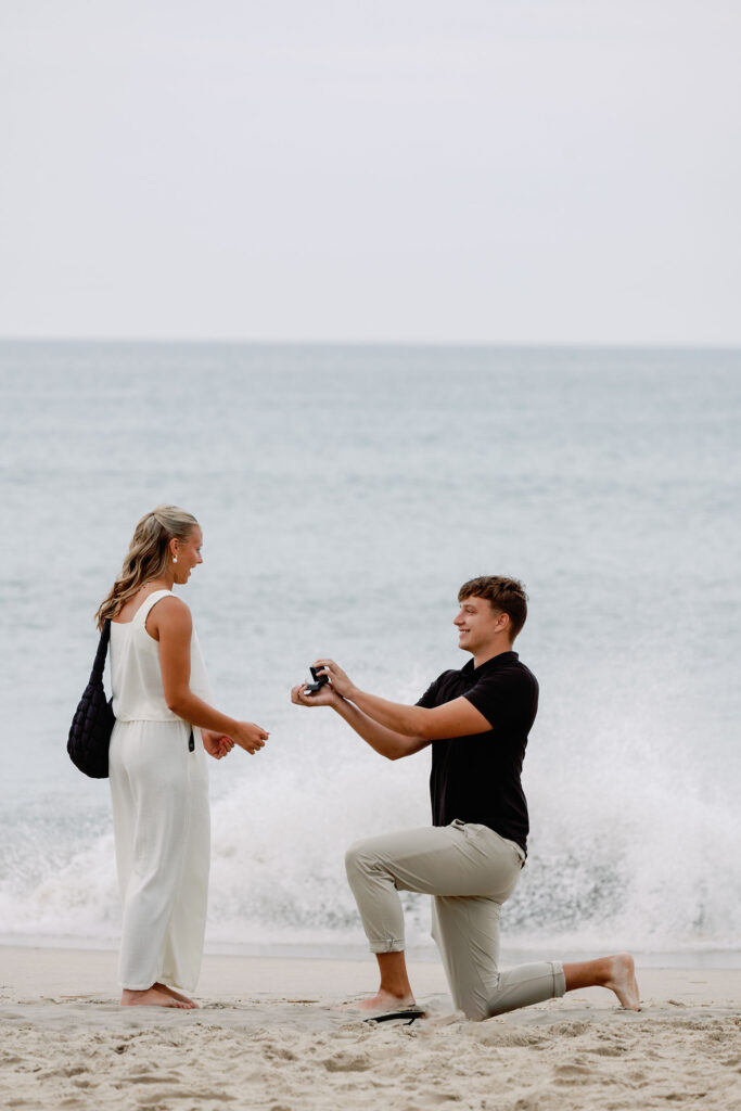 Groom-to-be kneeling in the sand during a surprise beach proposal at sunset on Emerald Isle, North Carolina.