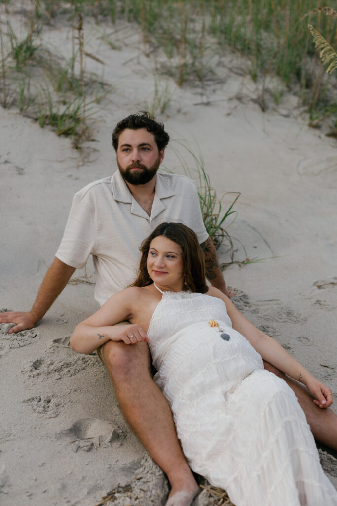 Intimate moment between couple seated on dunes, ocean breeze moving through tall grass.