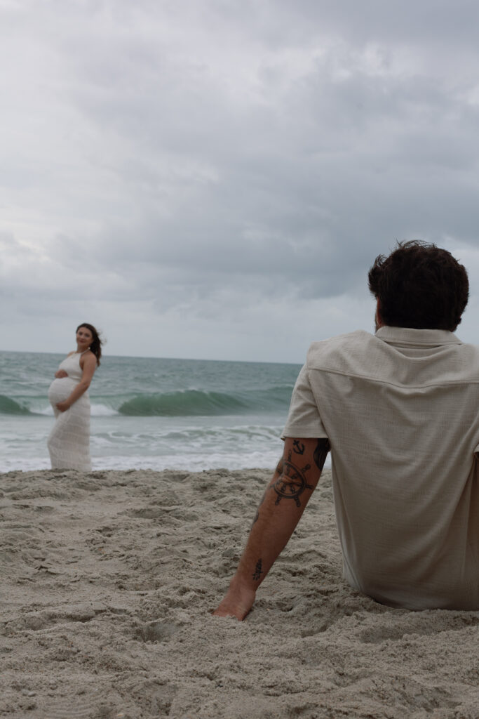 Partner sitting behind her on the dunes, both wrapped in quiet stillness as waves crash below.