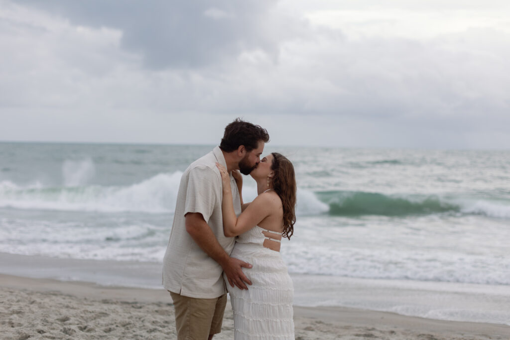 Final wide image of the couple looking toward the stormy horizon, the ocean reflecting muted color and calm.