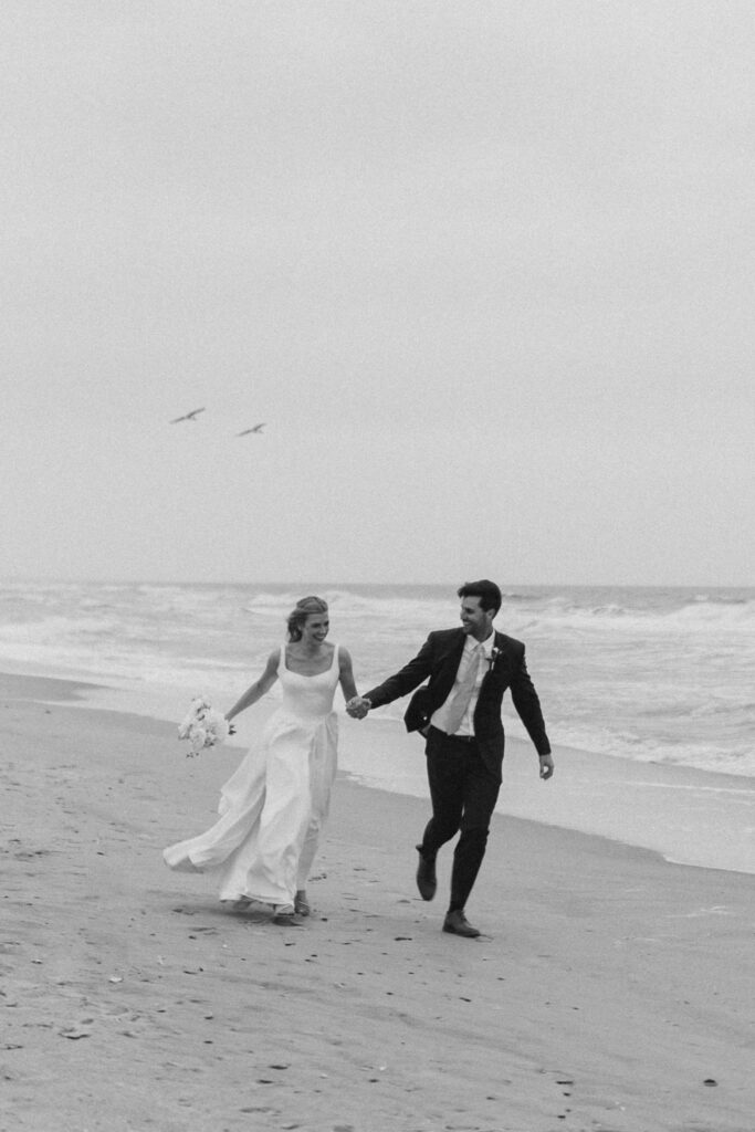 Bride and groom running hand in hand on the beach at The Dunes Club, Atlantic Beach, NC, black and white photograph by Hailey Rae Photography.