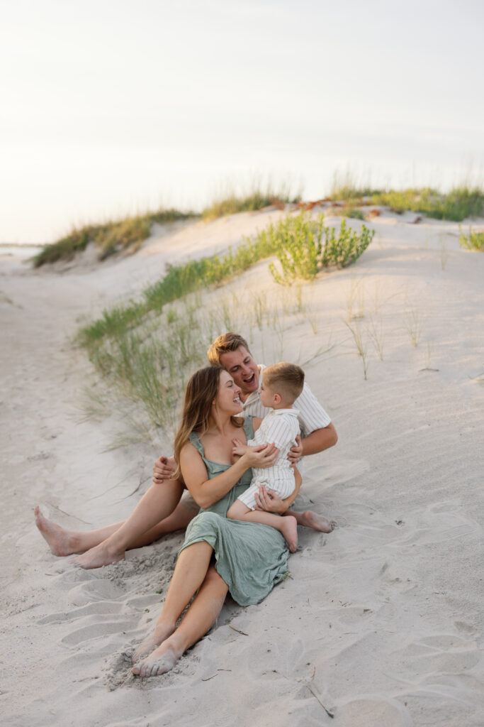 Candid family photography session in Atlantic Beach NC with parents and child cuddling on the sandy dunes at sunset.