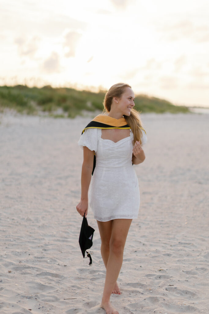 Graduate in cap and gown walking along the beach during her Crystal Coast senior session